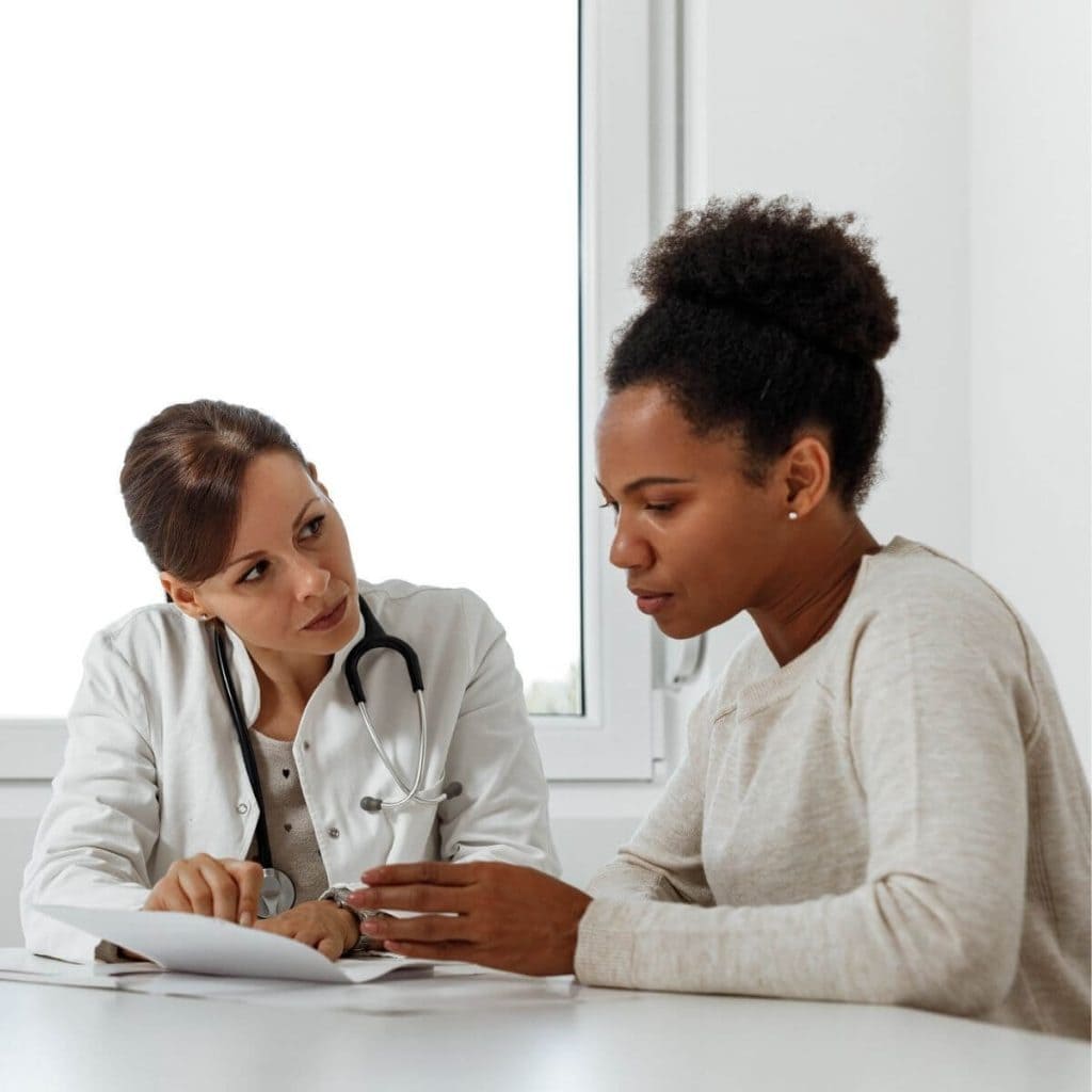 African American woman wearing a tan long sleeve shirt sitting at a table looking at a paper with a Mexican American female doctor wearing a white coat and stethascope