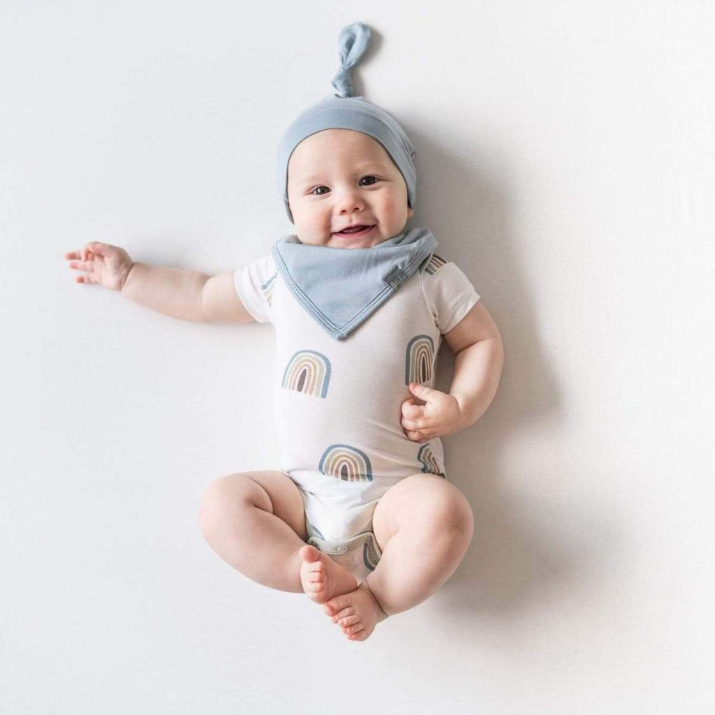 Baby in a white onesie with rainbows on it and a blue bib and knotted hat smiles on a white backdrop