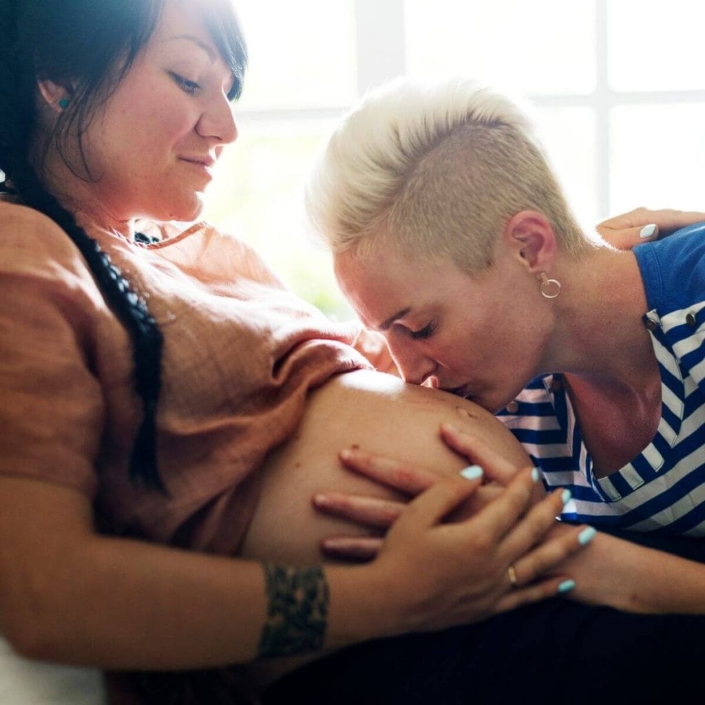 Woman with black hair in a braid with an orange gingham shirt pulled up over her pregnant belly looking down at a woman with very short blonde hair wearing a blue and white striped shirt with hands on the other woman's belly kissing it