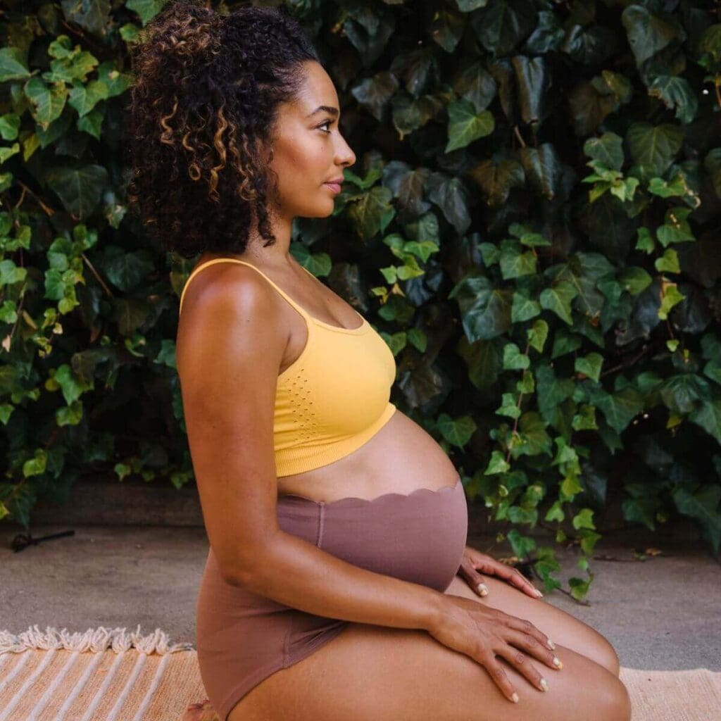 An African American woman with soft curly hair sits on her knees, facing sideways, with her hands on her thighs. She's wearing a yellow bralette and brown postpartum support bloomers from bao bei body