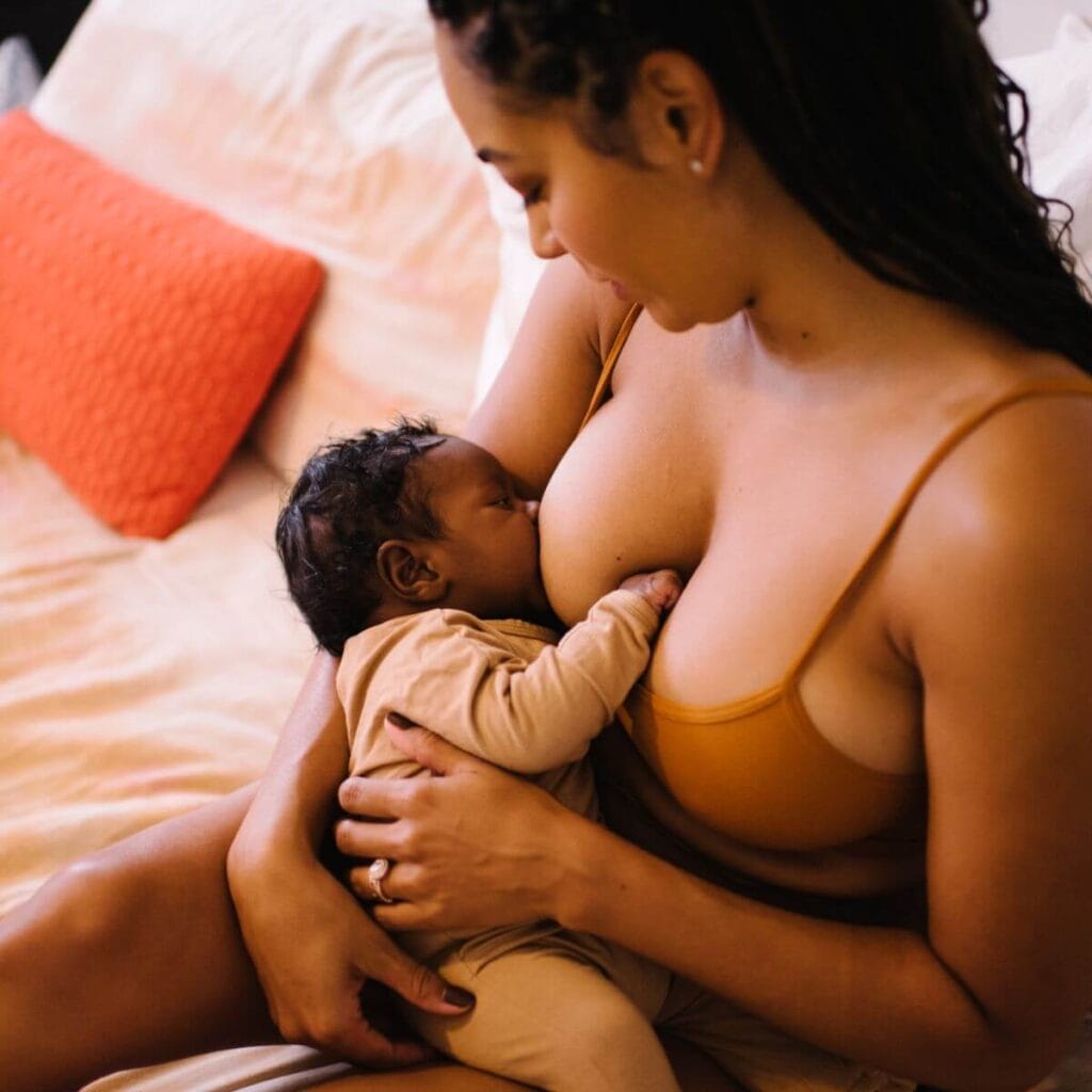a woman with golden brown skin and long dark curly hair rests on a bed wearing a brown nursing bra while breastfeeding a beautiful little curly-haired baby