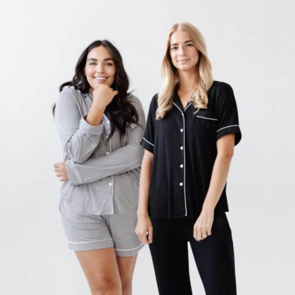 two women posing happily one with long dark hair wearing gray pajama shorts and a matching long-sleeved button-up pajama top. Her elbow is perched playfully on her hand, while her blonde friend stands next to her in long-sleeved black pajama pants with a matching short-sleeved button-up top. 