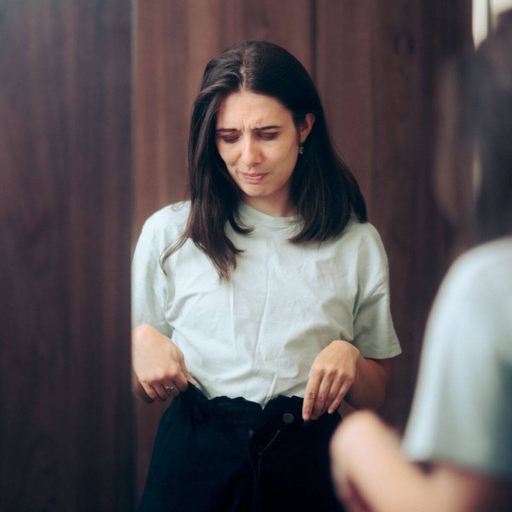 Caucasian woman with shoulder length dark brown hair wearing a white teeshirt and black pants holding the waist of the pants because she can't button them