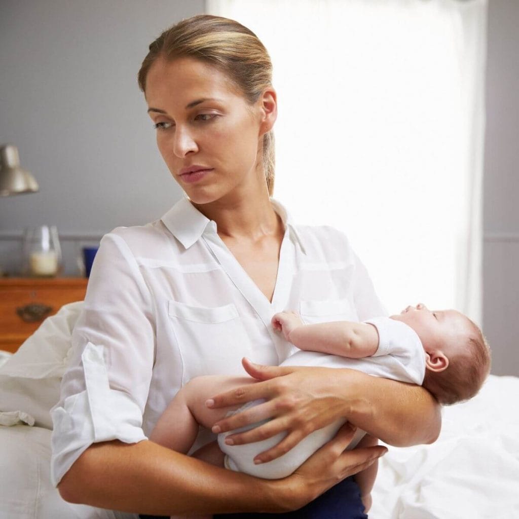 Caucasian woman sitting on a bed with white sheets wearing navy blue slacks and a white button up shirt looking sad while holding a sleeping baby in a short sleeve white onesie in her arms