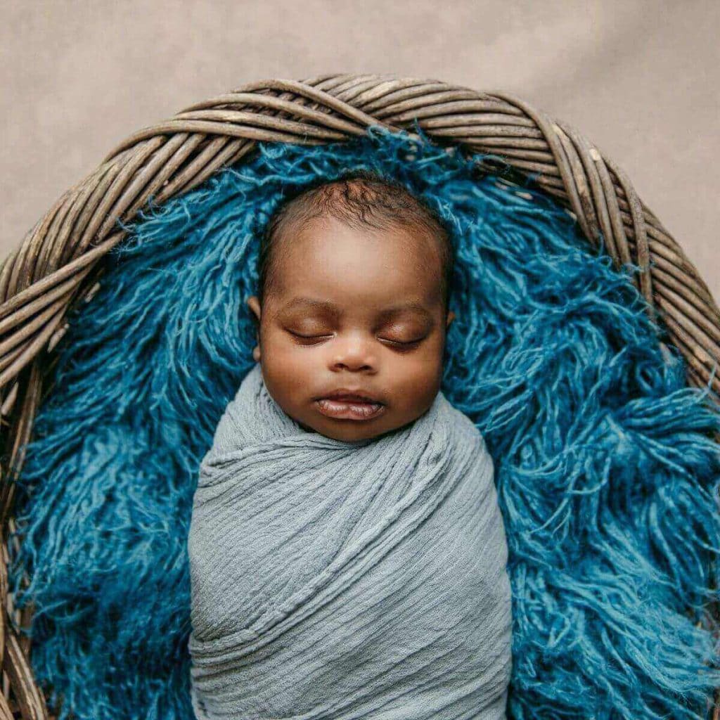 African American Newborn Baby Boy Asleep swaddled in a light blue blanket laying on top of bright blue yarn in a wicker basket