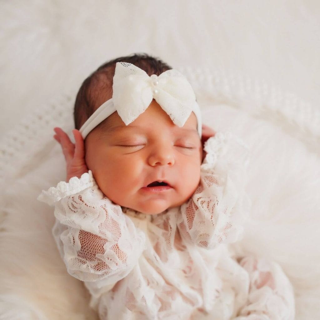 A newborn caucasian girl with her eyes closed and her hands on the sides of her face wearing a white lace outfit with large while bow headband laying on a white blanket