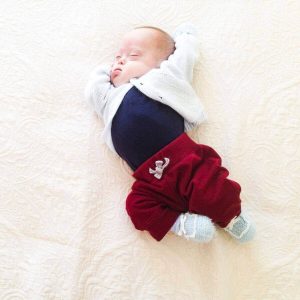 Newborn Caucasian baby with eyes close and hands under head wearing a white button up sweater, navy blue onesie, and maroon sweatpants with light blue and white booties laying on an all white blanket