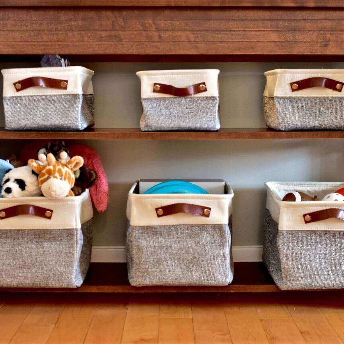 Six light grey and white cloth baskets with dark brown handles sitting on two wooden shelves filled with toys