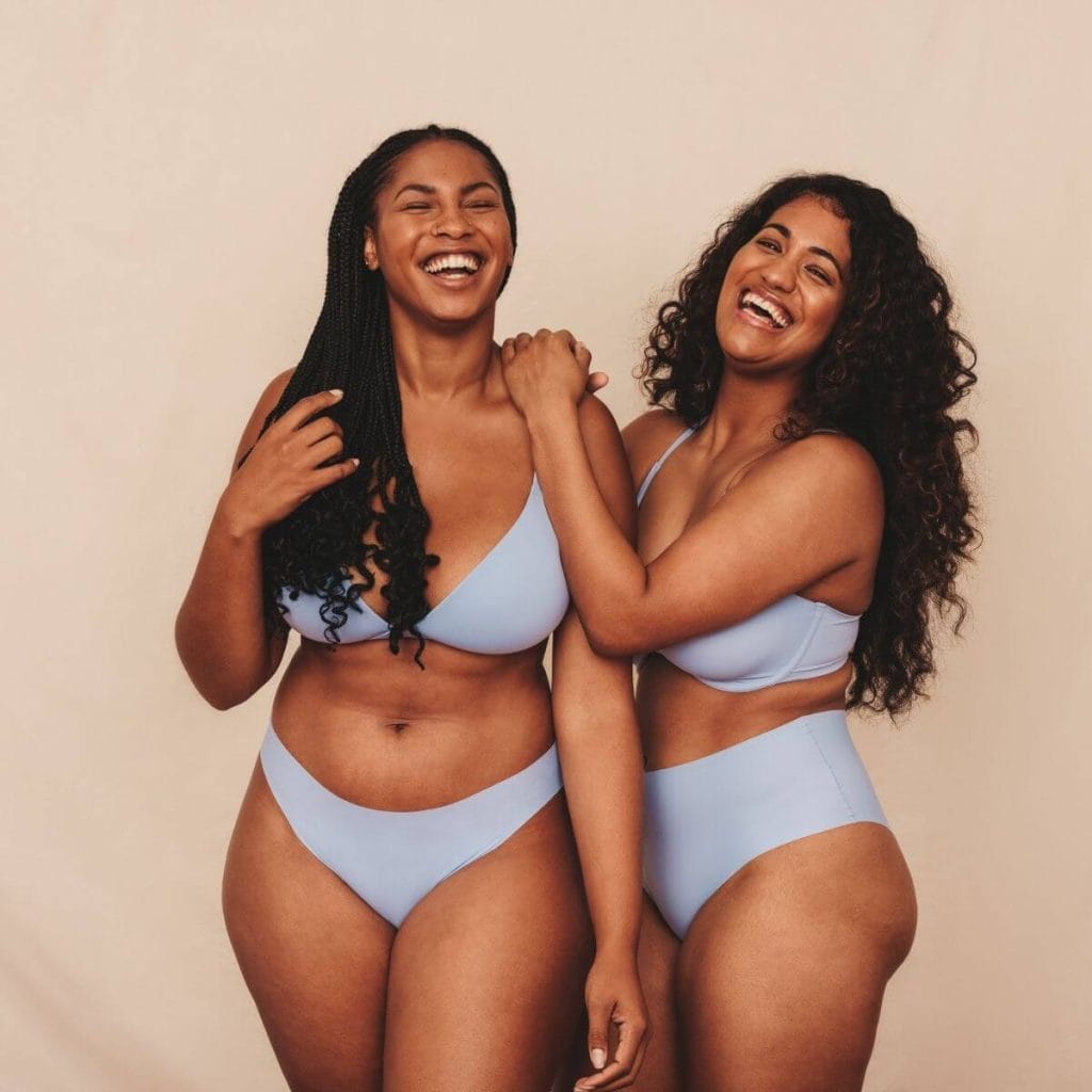 Two African American Women standing stide by side laughing both with long black hair wearing light blue bras and underwear