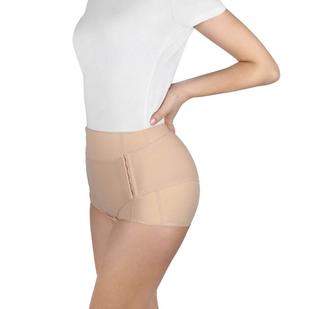 The middle section of a Caucasian woman's body with hands on back wearing a white t-shirt and tan postpartum support underwear