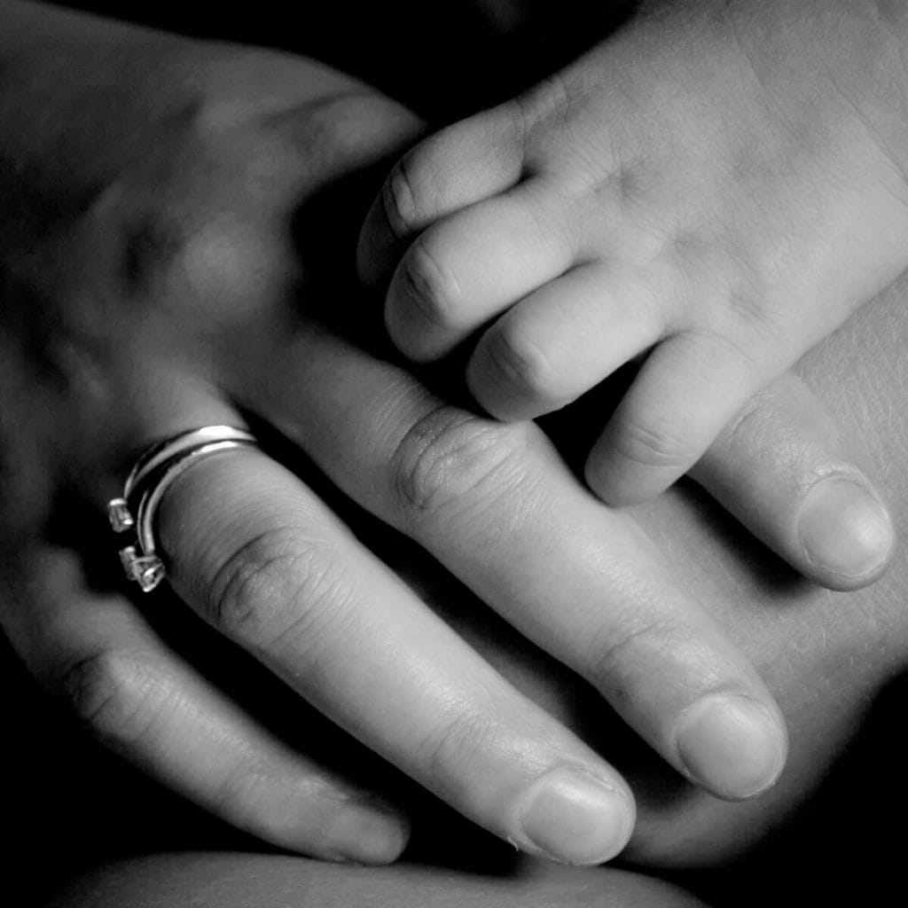Woman's hand with a heart ring on it with a newborn's hand gripping the pointer finger of the mother's hand