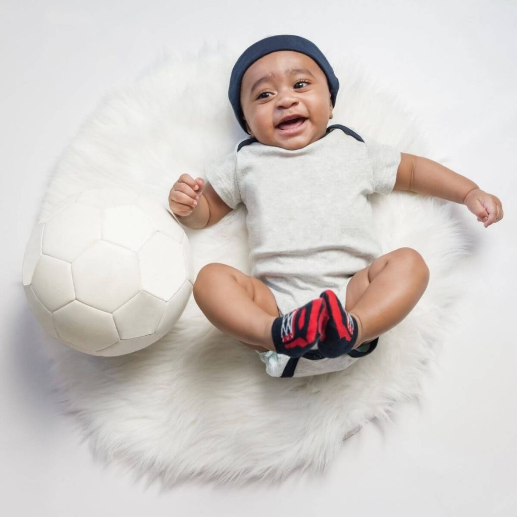 African American boy laying on a white fluffy round carpet wearing a dark grey hat, a white onesie with dark grey trim, and black and red socks that look like tennis shoes with a white soccer ball beside him