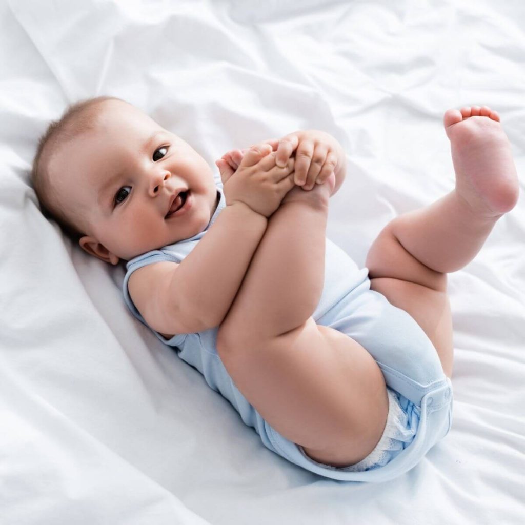 Caucasian baby staring at the camera laying on a white sheet with a light blue onesie on and holding their right foot in their hands.