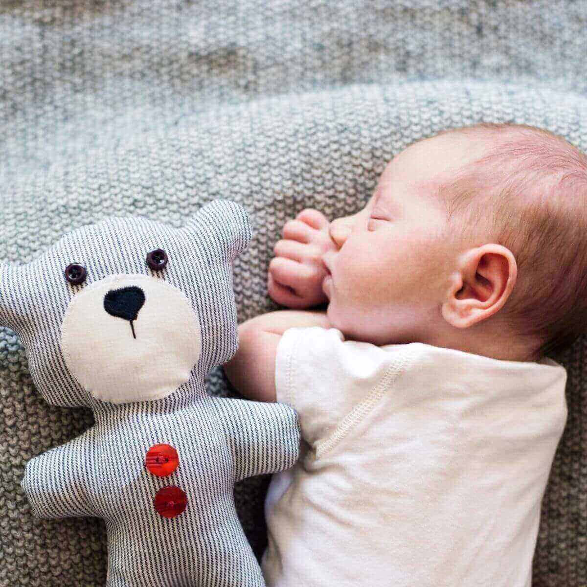 Newborn baby with eyes closed laying on stomach with a white onesie on. A light grey and white blanket is underneath the baby and a blue and white striped bear with red buttons is laying next to the baby