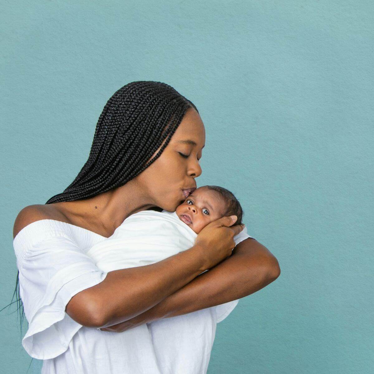 African American woman against a light blue background wearing a white shirt and kissing the heads of a newborn baby wrapped in a white blanket.