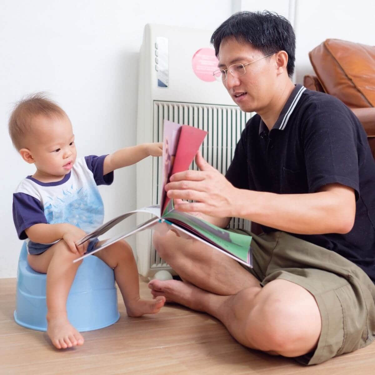 An Asian American dad sits on a wood floor with a book in his hand while his son is sitting on a light blue potty pointing to something in the book.