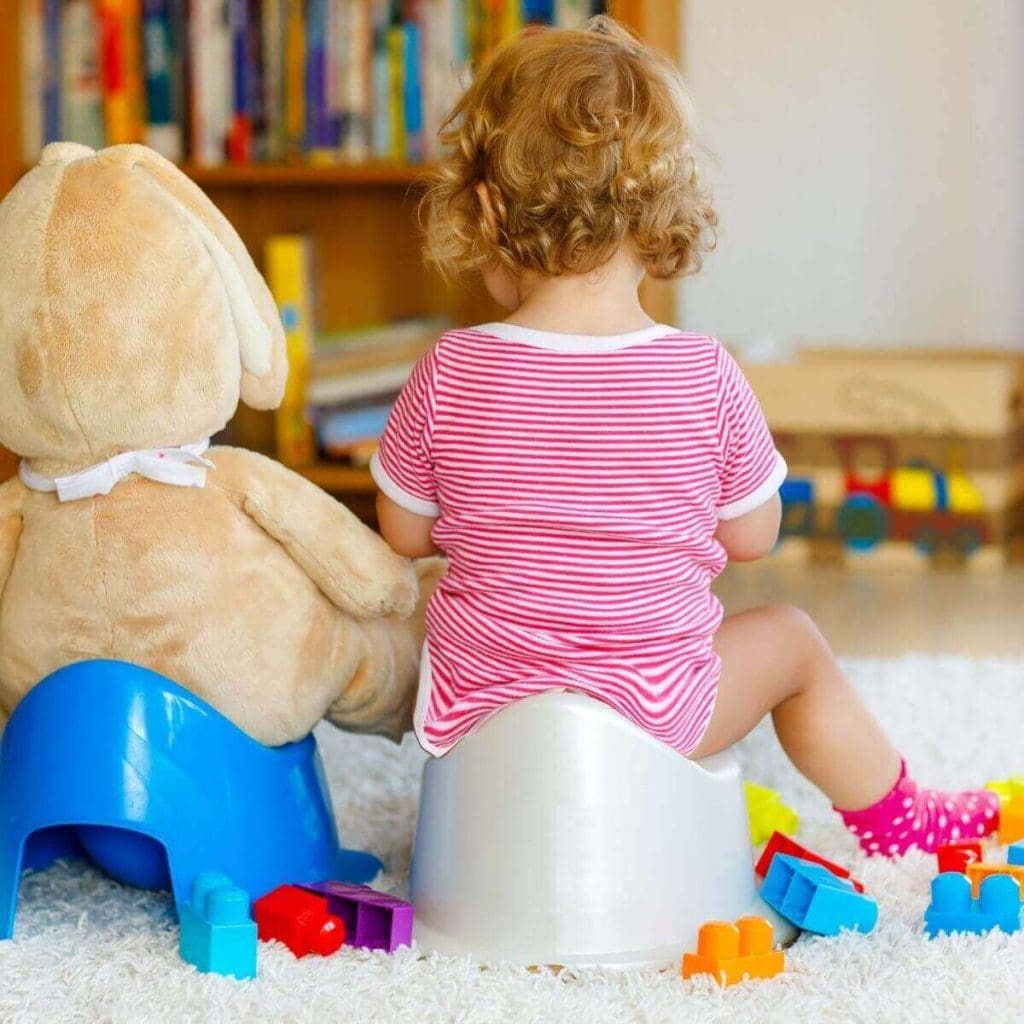 Little red headed girl in a pink and white striped onesie with her back to the camera sitting on a white potty. A light brown bear sits to her left on a blue potty. There is a bookcase and some toys in the background.