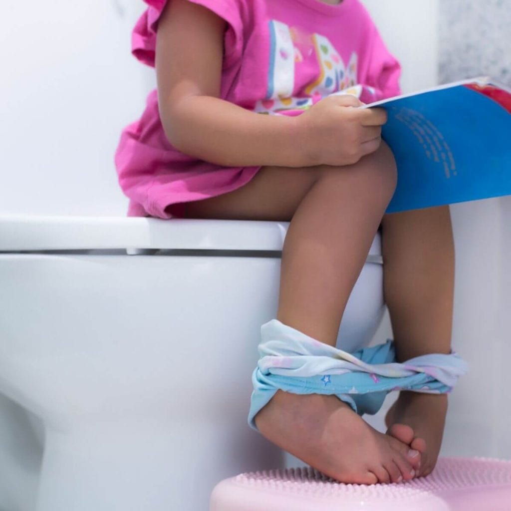 A little girl is sitting on a white toilet with a pink dress on and light blue underwear around her ankles holding a blue book with her feet on a light pink step stool.