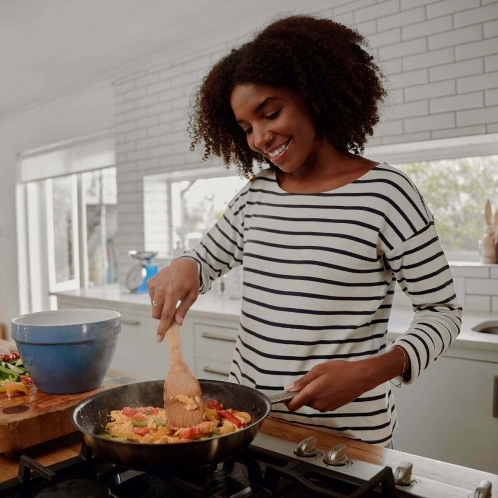 African American woman wearing a black and white striped long sleeve shirt standing in a large kitchen with white brick, large windows and white cabinets. She is cooking chicken and red, yellow, and green peppers in a frying pan and she is smiling.