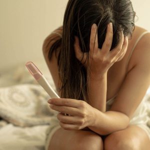 Woman with dark brown hair sitting on the side of a bed wearing a cream colored nighgown. Her hair is hiding part of her face and her hand is covering the rest of her face with a pregnancy test in her other hand.