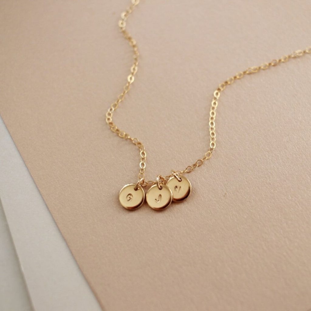 A thin gold link necklace chain is laying against a light pink background with three gold circles hanging from it with the letters "G", "J", and "M" initials