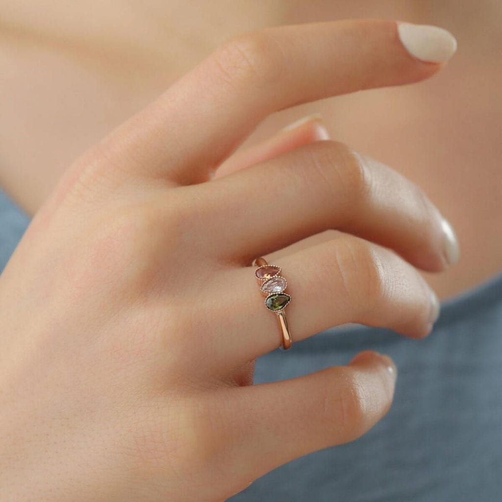 Woman's hand with white nail polish. On her ring finger there is a thin gold ring with 3 teardrop shaped birthstones on it.
