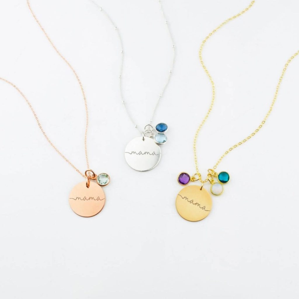 Three necklaces lay on a white table. One is a thin rose gold chain with a rose gold circle hanging from it that says Mama with a diamond birthstone, another is a silver chain with silver circle that says Mama with light and dark blue birthstones, and the last is a thin gold chain with a gold cirlce hanging from it that says Mama with opal, dark purple, and teal birthstones.