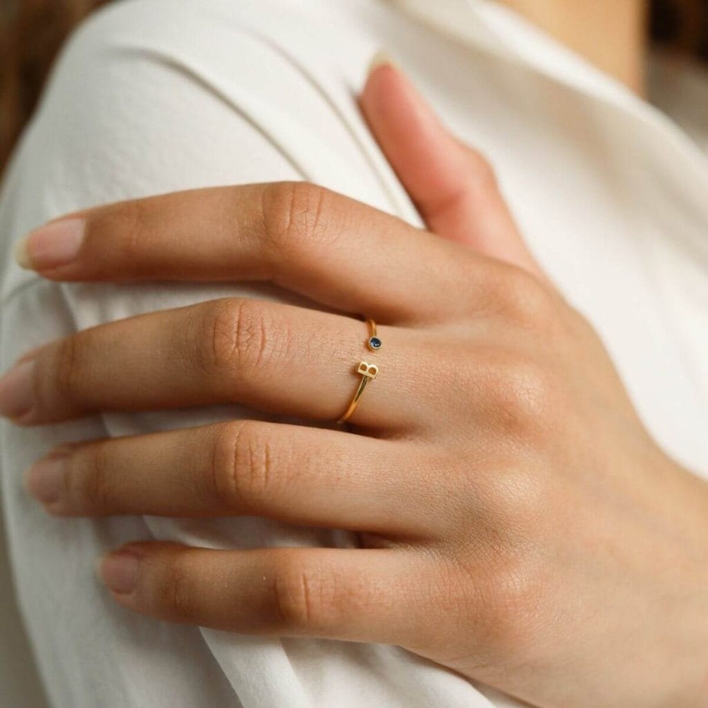 A woman's hand lays on the top of her arm with a thin gold ring with the letter B and a green birthstone