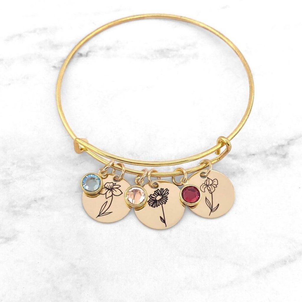A gold bracelet with 6 charms on it. Three of the charms are circles with birthstone flowers on them. The other three are circles with birthstones in them, a light blue stone, a clear stone, and a ruby stone