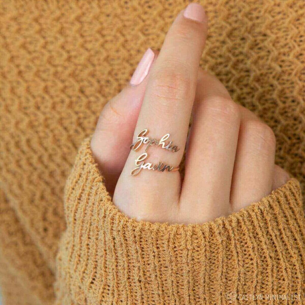 A woman wearing a yellow sweater holding up her hand with light pink fingernails with a gold ring on her pointer finger with the names Sophia and Gavin on it.