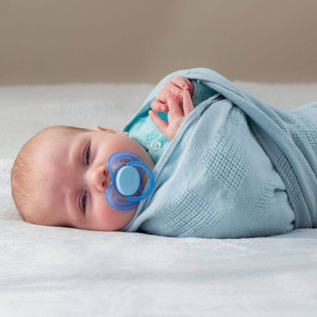Baby boy laying on a white sheet almost asleep with a blue pacifier in his mouth and wrapped in a blue blanket