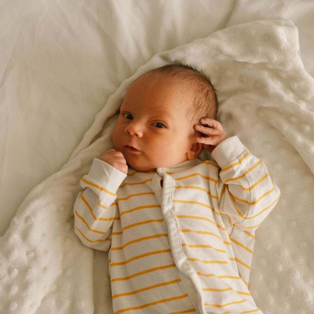 Newborn baby lays on a white blanket with puffy dots on it wearing a white and yellow striped sleeper waiting to be wrapped up
