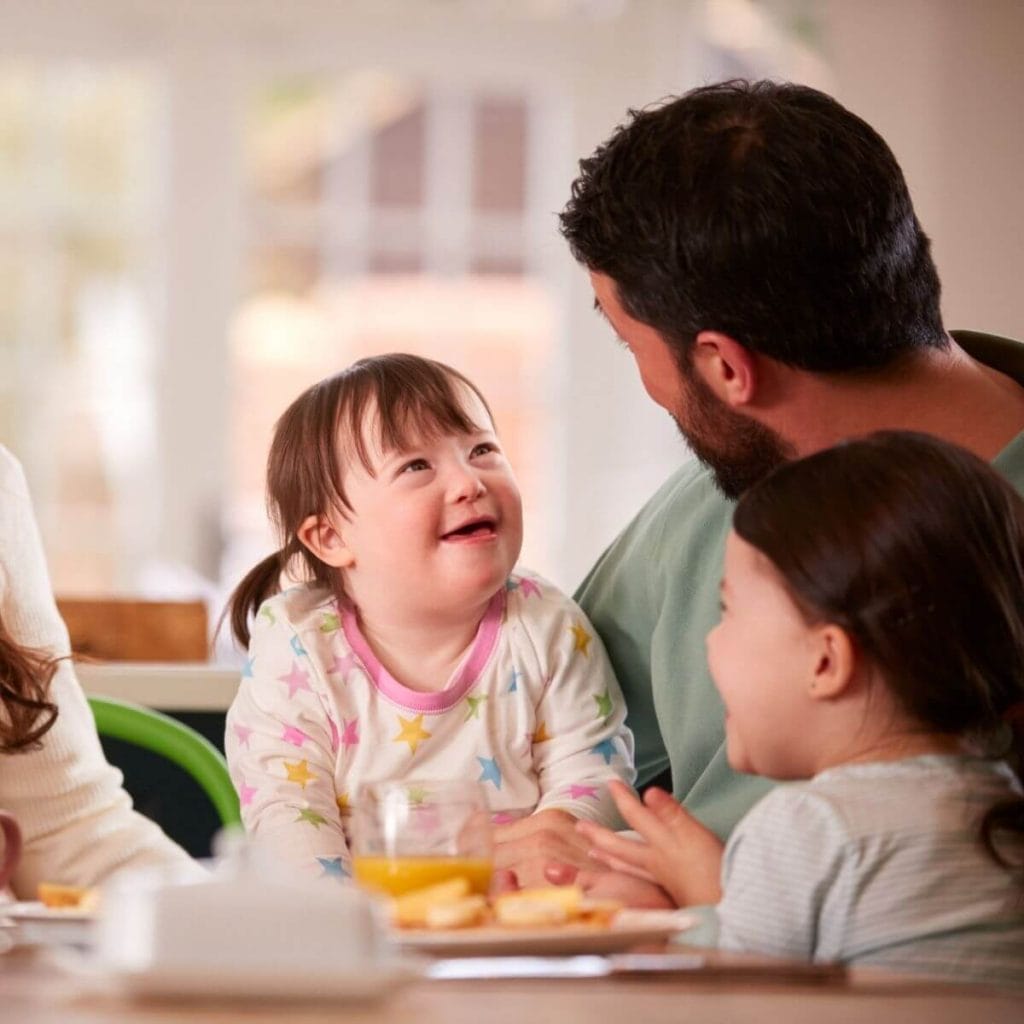 A father wearing a light green pajama shirt is sitting with his two daughters at the breakfast table. One daughter has on pajamas with pink, yellow, green, and blue stars and the other is wearing blue and white striped pajamas.