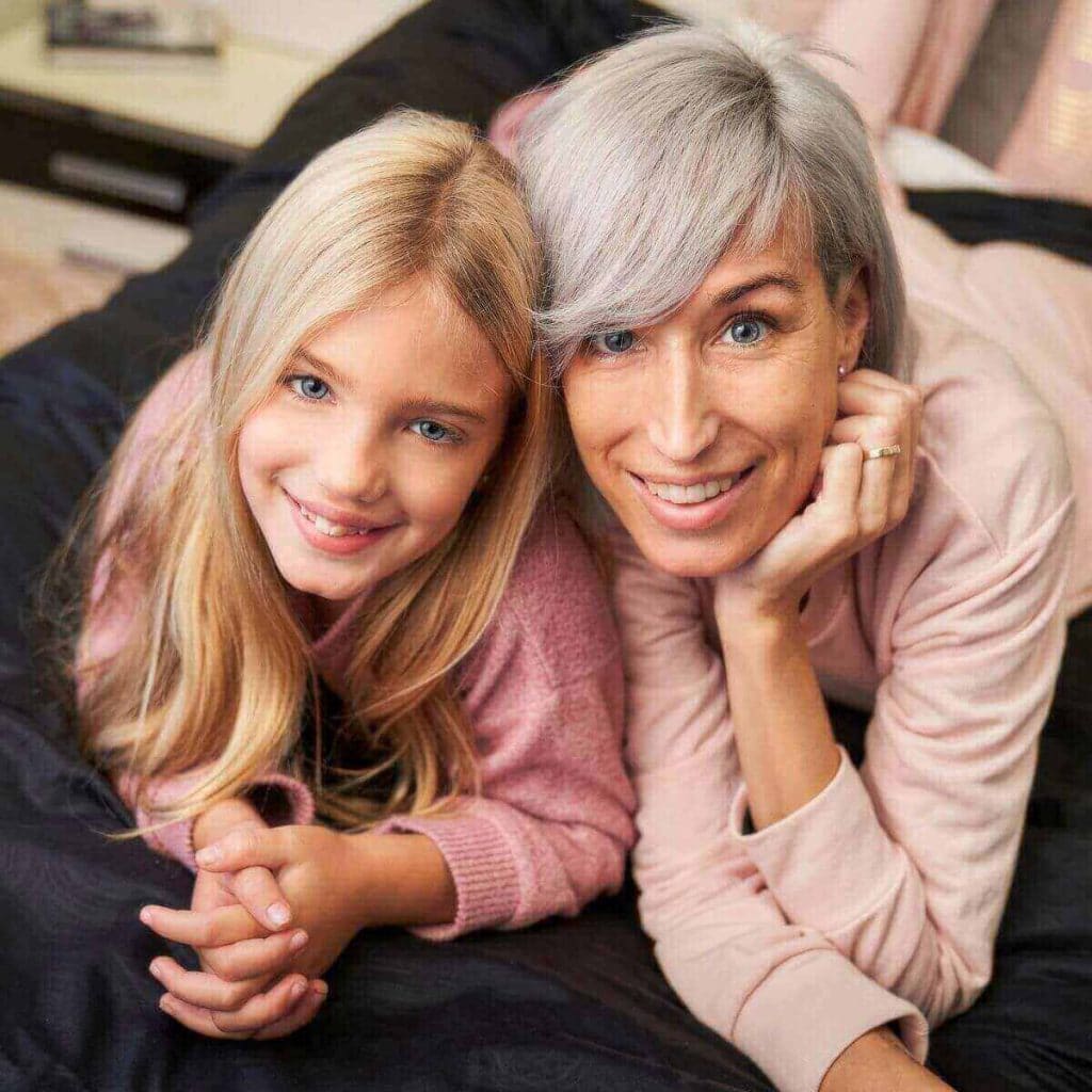 A woman with grey hair wearing light pink pajamas is laying on a black blanket beside her daughter who is wearing dark pink pajamas. Both are smiling at the camera.