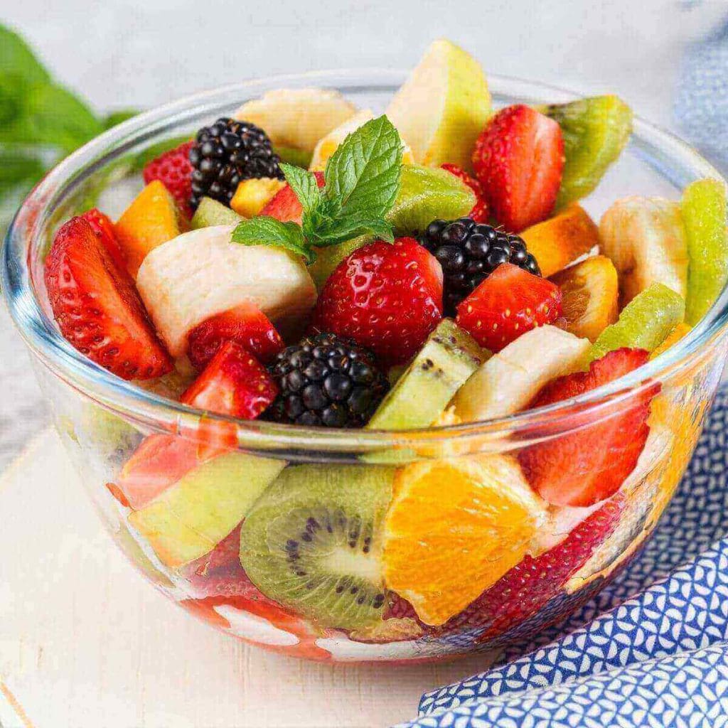 A glass bowl sits on a white table with a blue and white napkin. In the bowl is a mixture of bananas, strawberries, kiwi, oranges, blackberries, and green apples