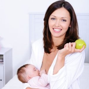 A Caucasian woman in white bathrobe breastfeeding a baby and holding a green apple