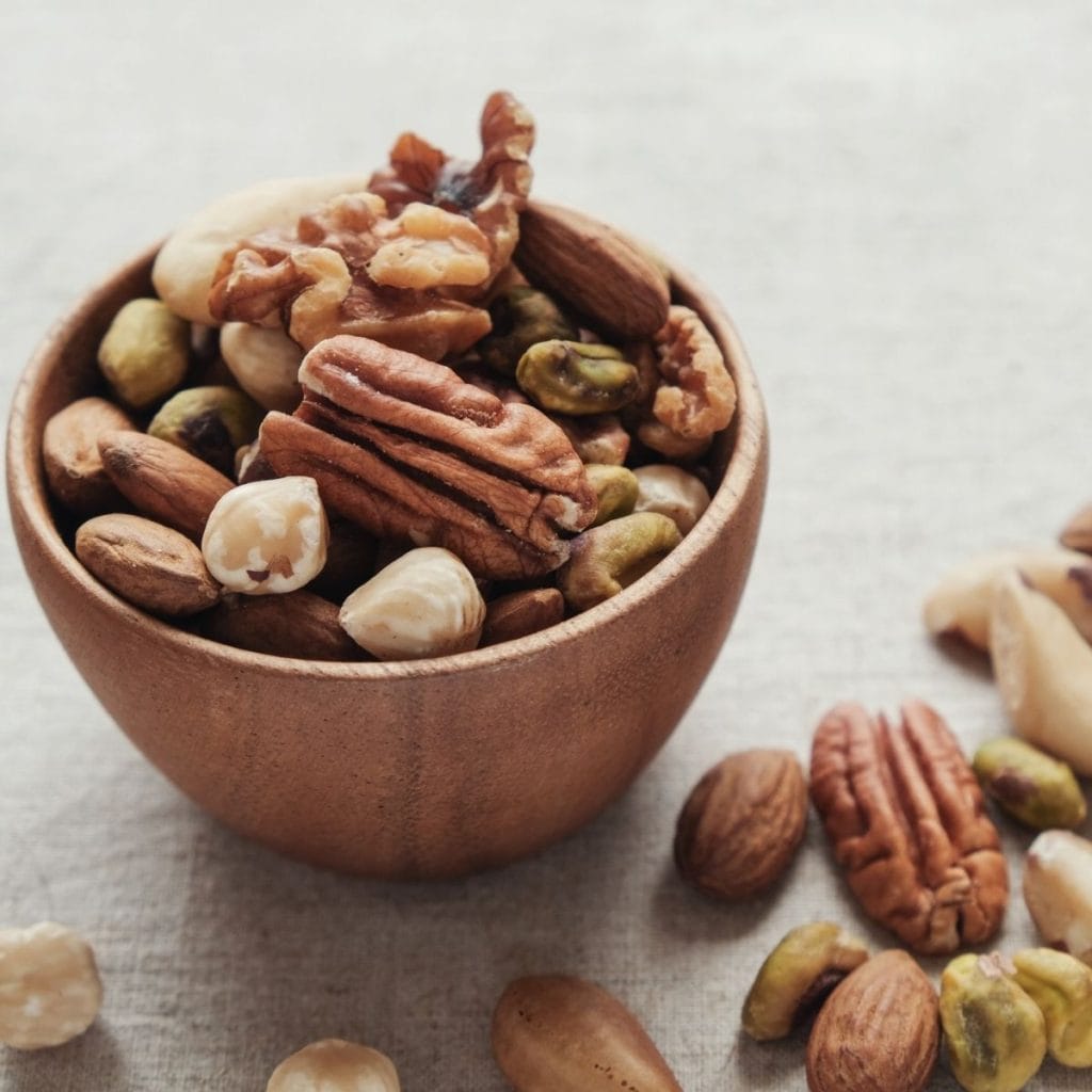 A dark wood bowl sits on a table with a light gingham tablecloth. In the bowl are many different types of nuts including cashews and pecans.