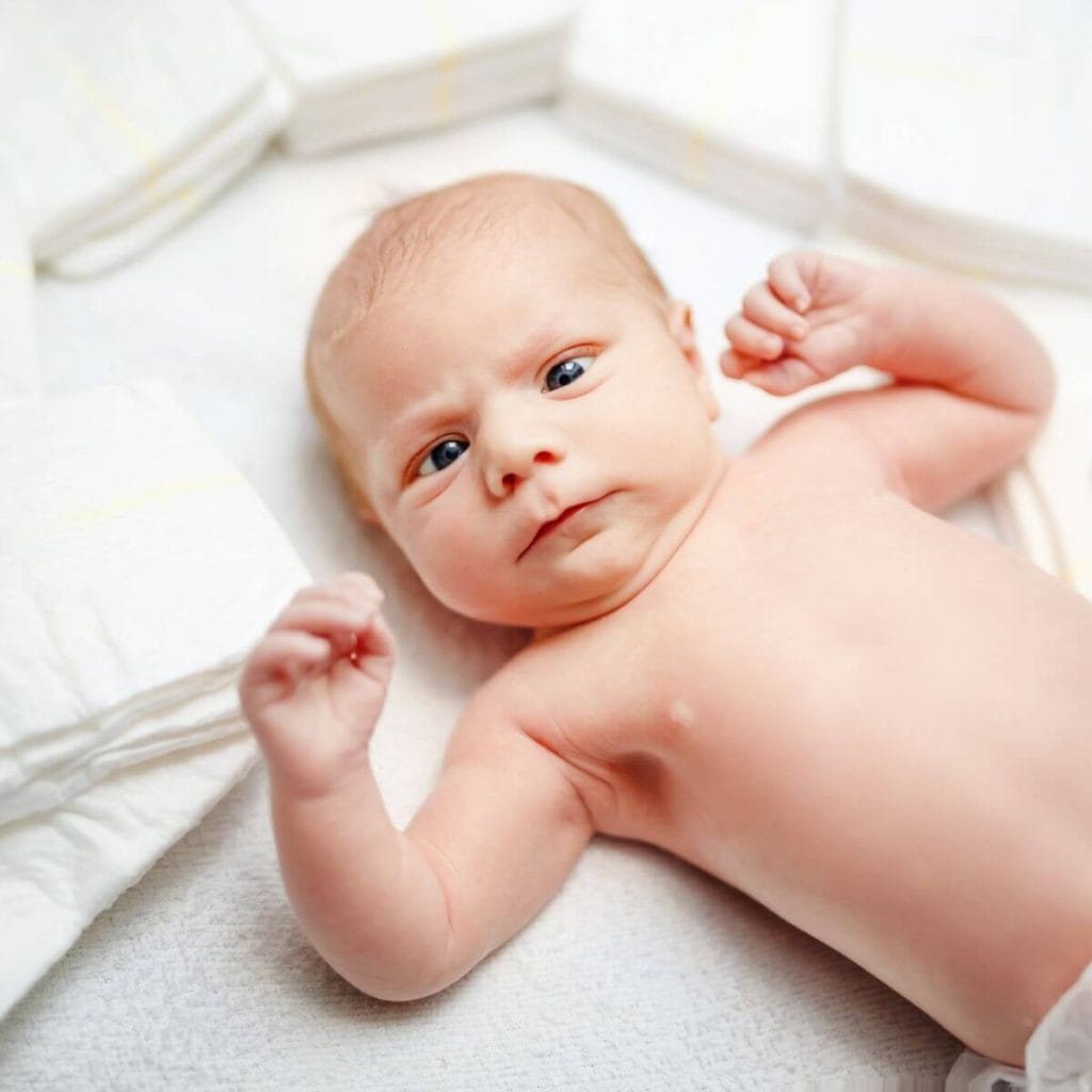 Caucasian baby is laying on a white blanket wearing nothing but a diaper. It's blue eyes are staring directly at the camera and there are stacks of diapers laying all around the baby.
