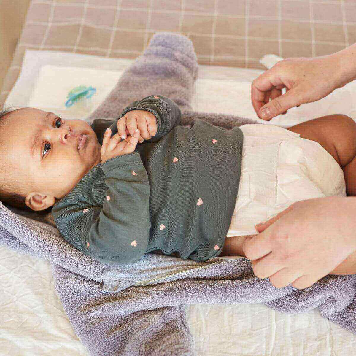 African American baby is laying on a puppy pad after getting their diaper changed. The baby is wearing a dark grey shirt with little light pink hearts all over it