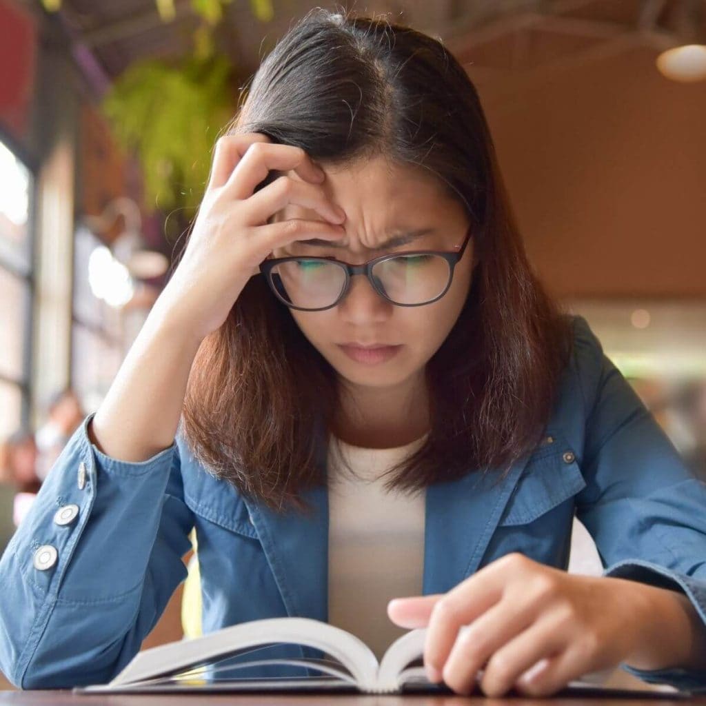 Asian American woman with shoulder length dark brown hair and wearing black rimmed glasses is sitting at a table in a coffeehouse with her right hand on her forehead as she reads a book that is laying on the table in front of her.
