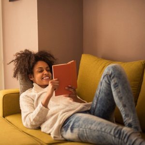 African American woman is laying down on a mustard yelow couch wearing a cream colored sweater and light blue jeans, smiling and reading an orange covered book