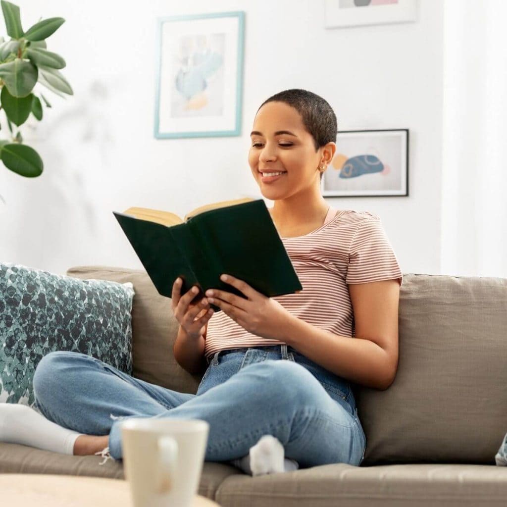 African American woman is sitting on a grey couch with her legs folded under her, wearing a red and white striped shirt and light blue jeans with white socks, laughing and holding a black covered book
