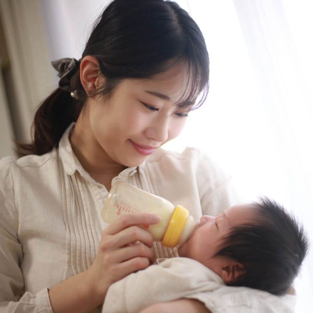 Asian American woman is standing beside a window with her hair pulled back in a low ponytail and wearing a long sleeve cream shirt smiling down at a baby in her arms that she is bottle feeding