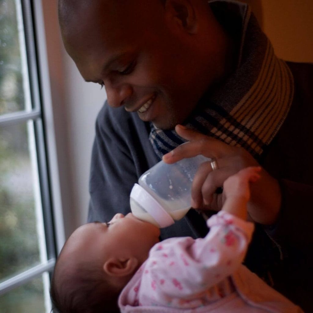 African American man wearing a dark grey shirt is standing by a window and smiling down at the newborn girl in his arms while he bottle feeds her