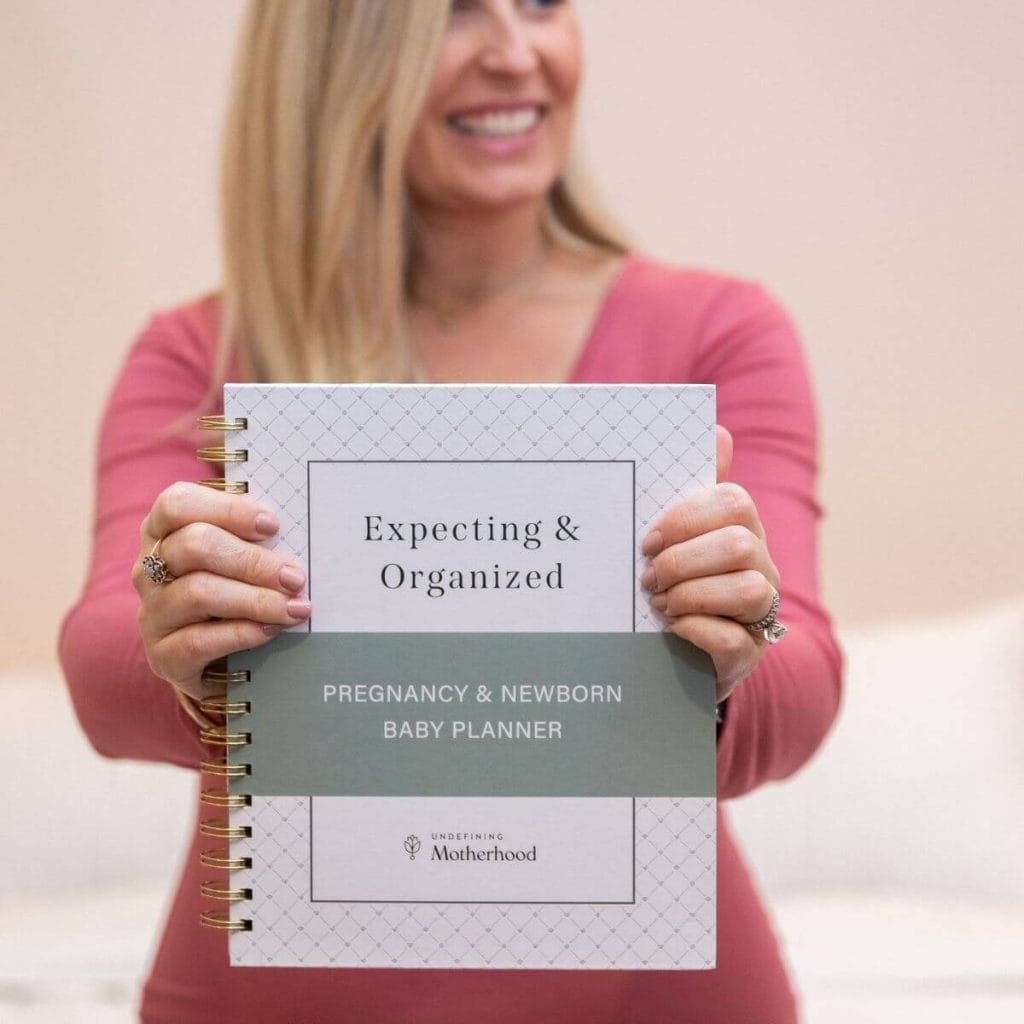 Blonde woman in pink dress holding spiral bound pregnancy planner that says expecting and organized pregnancy and newborn baby planner on the cover