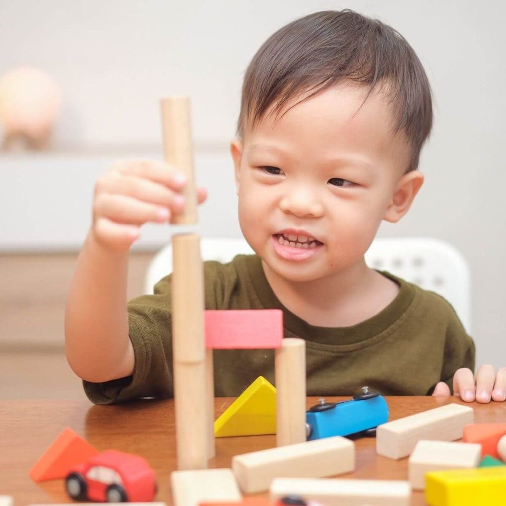 An Asian American toddler boy is sitting at a light wood colored table in a white chair. He is wearing an olive green short sleeve t-shirt and is smiling while stacking wooden cylinders