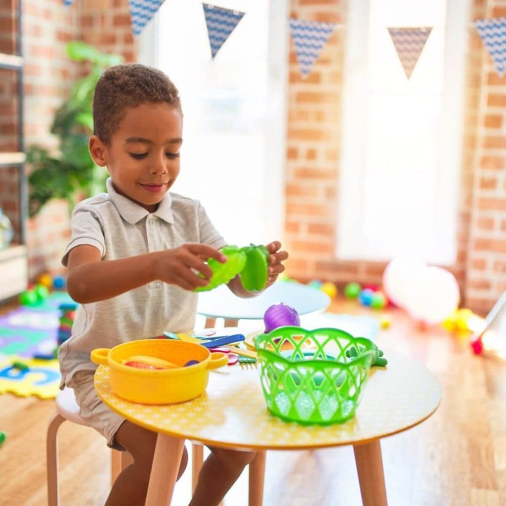 An African American toddler boy is wearing light kakhi shorts and a light grey short sleeve shirt with a collar. He is sitting at a yellow and white checkered table with a light green basked, a yellow pot with two handles and play food and utensils around him. He is holding a green pepper in his hands