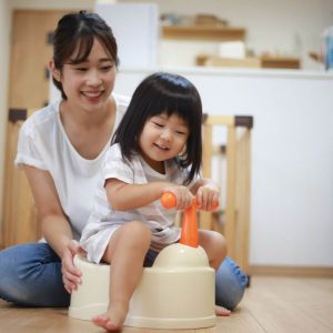 An Asian American mom sits on a wood floor in the living room while her daughter is sitting on a cream potty training seat with an orange handle in the front that she is holding onto.