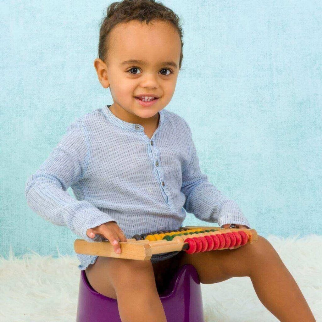 An African American boy is wearing a light blue long sleeve shirt while sitting on a purple potty training seat with a toy in his hand.