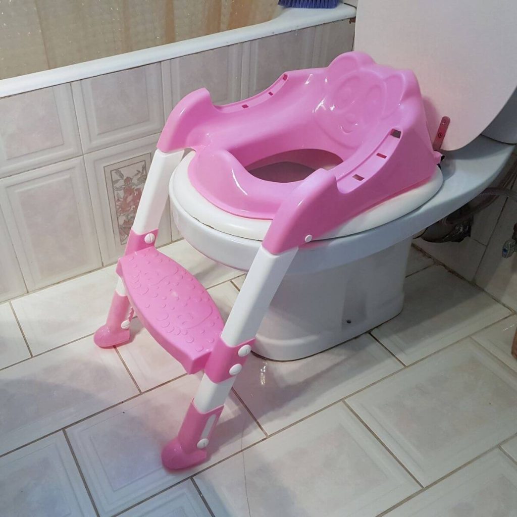A white toilet sits in a bathroom with light pink and white tile. There is a bright pink potty training seat on the toilet with a step ladder attached.